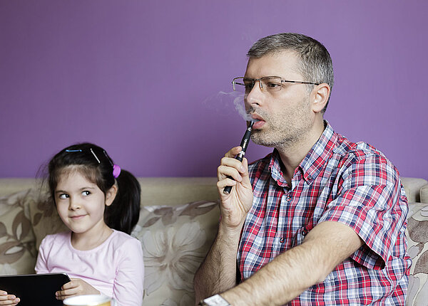 Erwachsener Mann sitzt auf einer Couch neben einem kleinen Mädchen und raucht eine E-Zigarette. Er trägt ein kurzes kariertes Hemd und das Mädchen hat ein Tablet in der Hand