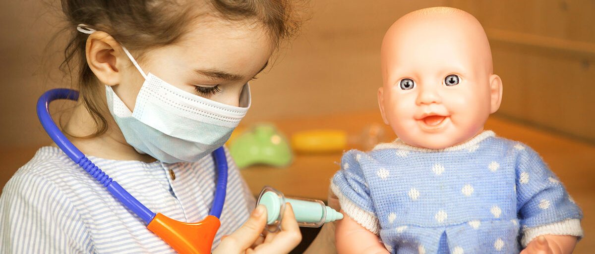 Ein kleines Mädchen trägt einen Mundschutz, um den Hals hat sie ein Plastik-Stethoskop hängen. Mit einer Spielzeug-Spritze gibt es einer Puppe eine Impfung in den Oberarm.