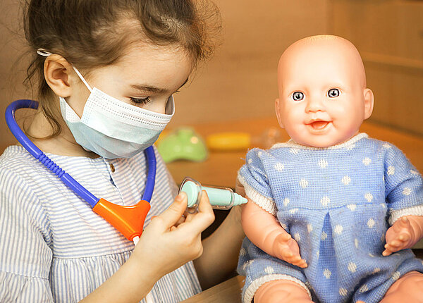 Ein kleines Mädchen trägt einen Mundschutz, um den Hals hat sie ein Plastik-Stethoskop hängen. Mit einer Spielzeug-Spritze gibt es einer Puppe eine Impfung in den Oberarm.