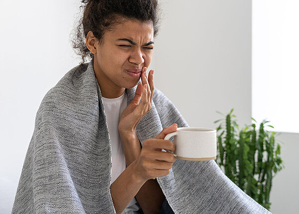 Zahnschmerzen bei Erkältung Junge afroamerikanische Frau hat eine Decke über den Schultern, hält eine Tasse in der Hand und hält sich mit der anderen Hand schmerzverzerrt den Mund.