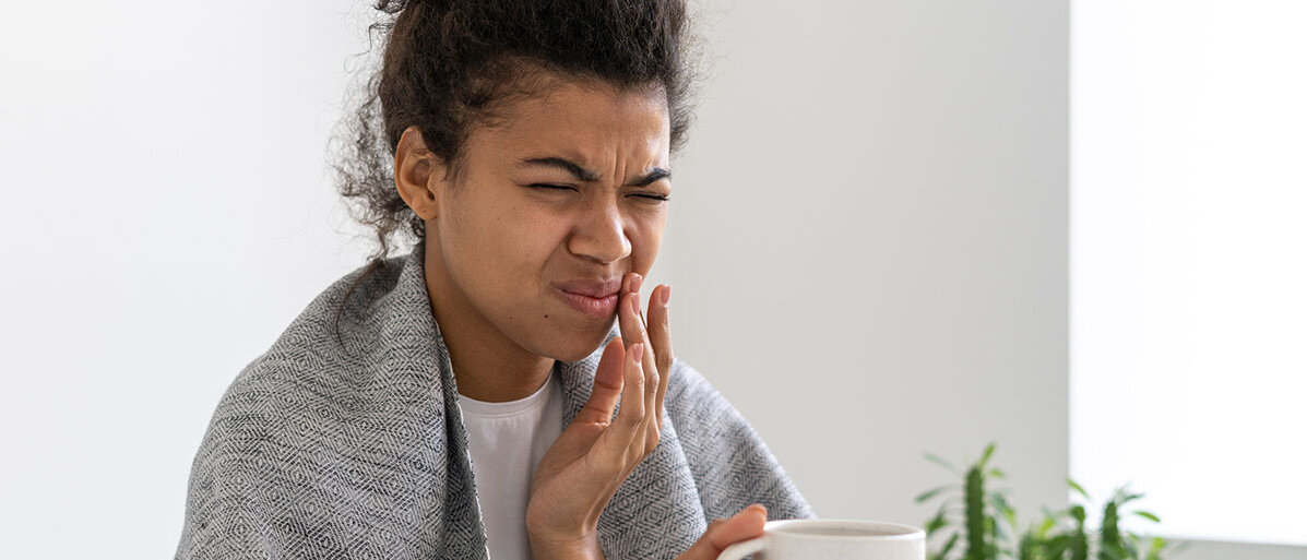 Junge afroamerikanische Frau hat eine Decke über den Schultern, hält eine Tasse in der Hand und hält sich mit der anderen Hand schmerzverzerrt den Mund.