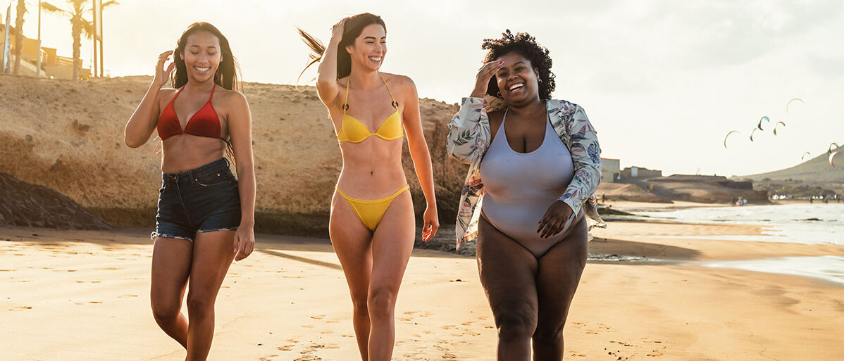 Drei Frauen in Badebekleidung laufen lachend am Strand entlang.