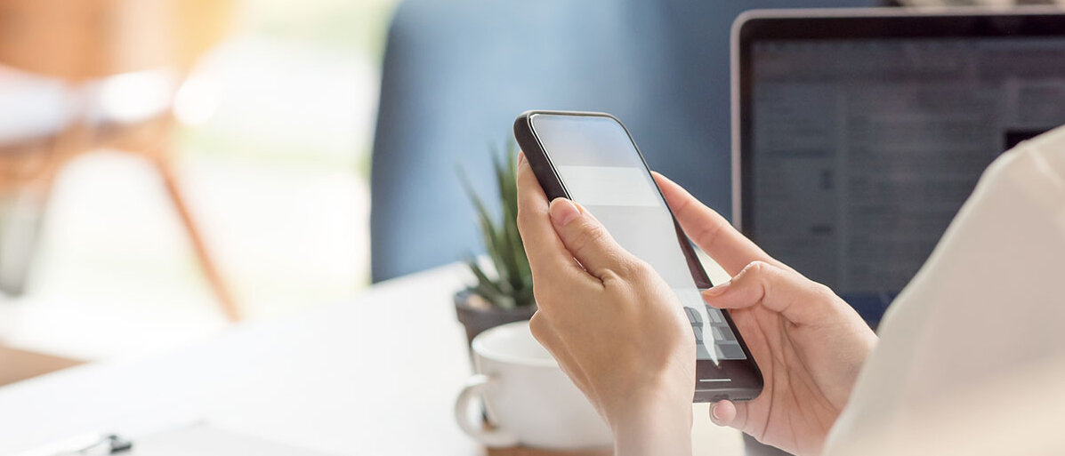Frau schaut auf ihr Handy und rechts nebendran ist ihr Laptop zu sehen