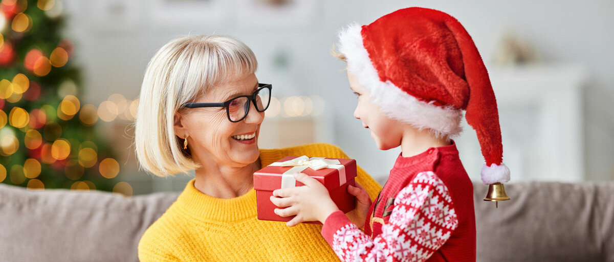 Eine Rentnerin sitzt auf dem Sofa und hält ihren Enkel im Vorschulalter auf dem Schoß. Er trägt eine rote Nikolausmütze mit Glöckchen an der Spitze, einen Weihnachtspullover und überreicht ihr ein rotes Geschenk. Im Hintergrund leuchtet der Weihnachtsbaum.