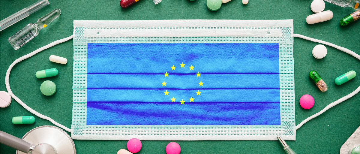 Ein Flatlay auf dunkelgrünem Hintergrund: Mehrere lose Tabletten und Kapseln, Tablettenblister, ein Fieberthermometer, Vials und ein Stethoskop liegen um eine chirurgische Maske herum drapiert. Die Maske zeigt die EU-Flagge.