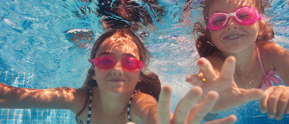Zwei Mädchen mit pinken Schwimmbrillen tauchen im Schwimmbad, strecken ihre Hände in Richtung der Kamera und lachen.
