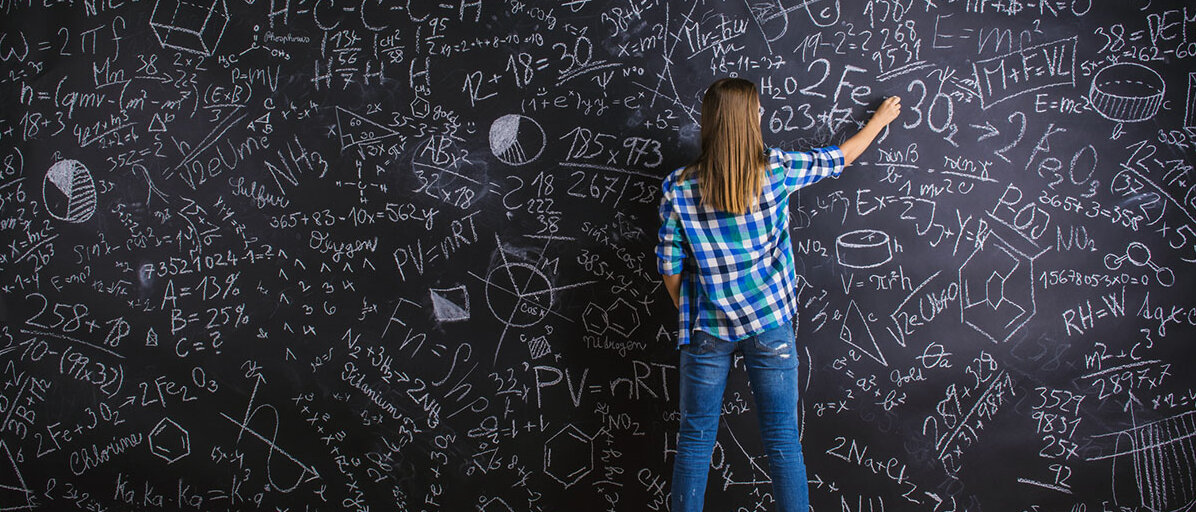 Eine Frau steht vor einer übermannshohen Tafel und schreibt sie voll mit wissenschaftlichen Formeln.
