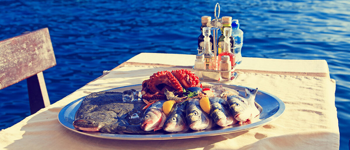 Übertragung und Krankheitsbild bei Hepatitis A und B Teller mit Meeresfrüchten auf einem Tisch und im Hintergrund das Meer
