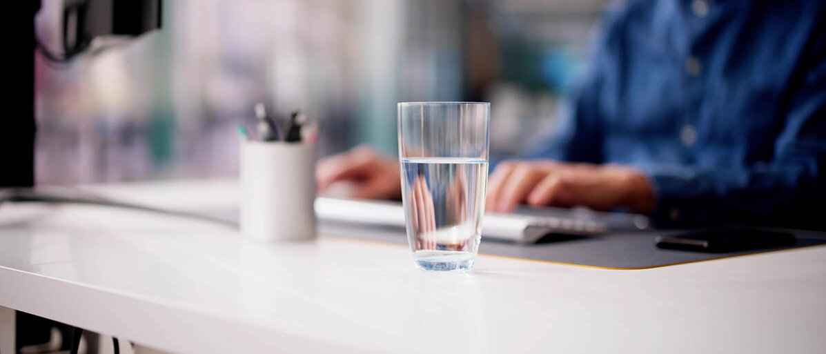 Im verschwommenen Hintergrund arbeitet ein Mann am Computer. Im Vordergrund steht deutlich ein unangetastetes Glas Wasser.