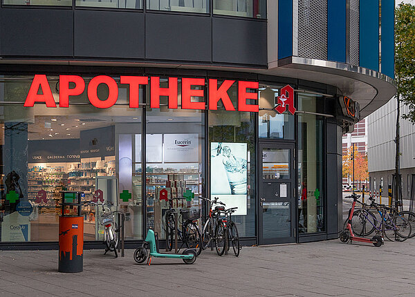 Eingang einer Apotheke mit Glasfassade. Davor stehen Fahrräder und Roller.