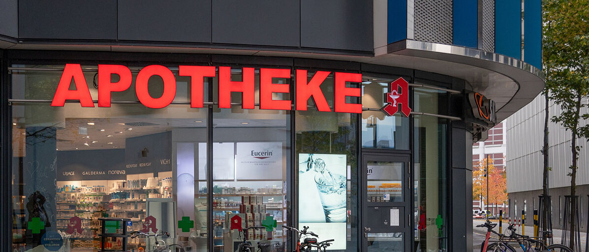 Apothekenvergütung und Apothekenreform Eingang einer Apotheke mit Glasfassade. Davor stehen Fahrräder und Roller.