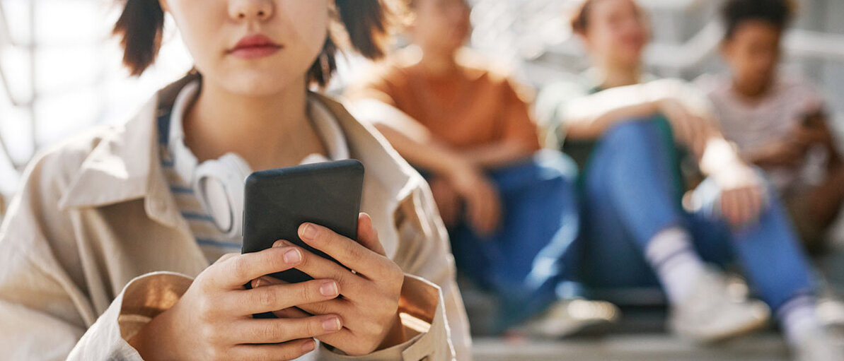 Mentale Gesundheit auf Social Media Gruppe von Jugendlichen sitzt auf einer Treppe. Im Vordergrund ein junges Mädchen, dass gedankenverloren auf ihr Handy blickt.