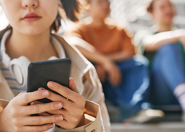 Mentale Gesundheit auf Social Media Gruppe von Jugendlichen sitzt auf einer Treppe. Im Vordergrund ein junges Mädchen, dass gedankenverloren auf ihr Handy blickt.