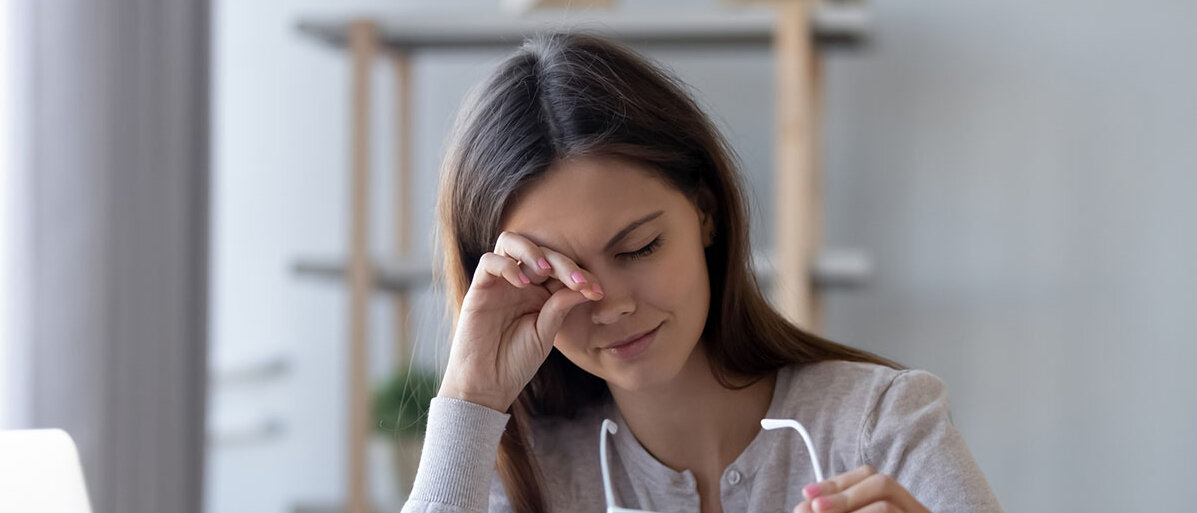 Trockene Augen im Winter Frau sitzt am Schreibtisch vor ihrem Laptop, reibt sich mit einer Hand ein Auge und mit der anderen hält sie ihre Brille fest.