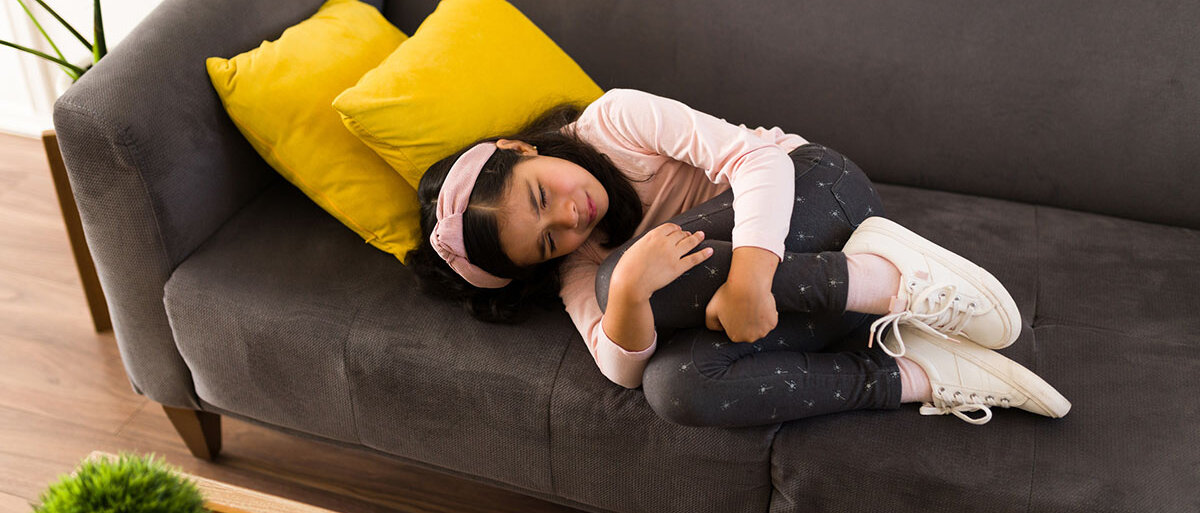 Ein Mädchen liegt zusammengekrümmt auf dem Sofa und verzieht das Gesicht vor Schmerz.