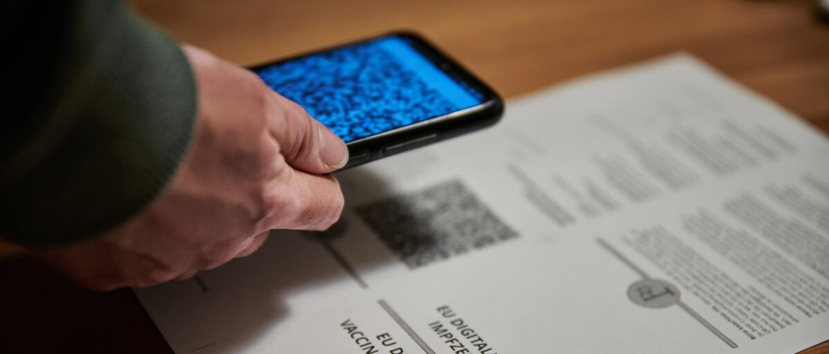 Ein Ausdruck mit QR-Code für das digitale Impfzertifikat liegt auf dem Tisch. Eine Hand hält ein Smartphone darüber und scannt den QR-Code ein.