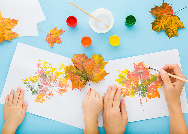 Ein Kind und eine Erwachsene malen ein herbstlich bunt gefärbtes Blatt nach, das sie vor sich auf dem Tisch liegen haben.
