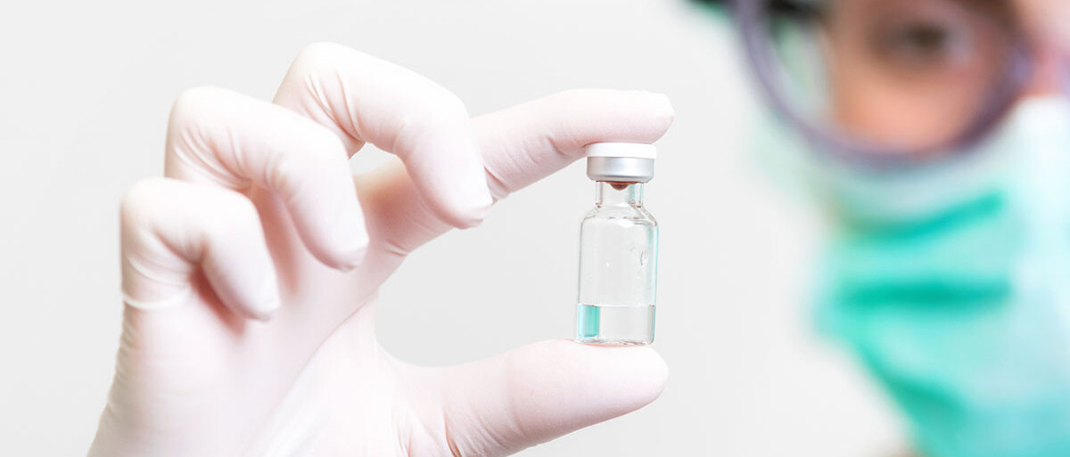 Eine Person in Schutzausrüstung hält zwischen Daumen und Zeigefinger ein Impfstoffvial und begutachtet es.