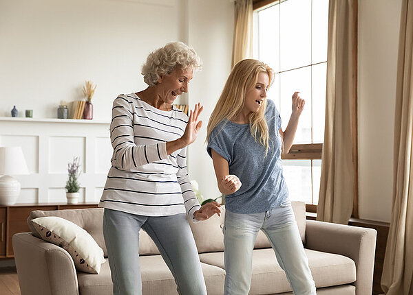 Zwei Frauen, eine in den 30ern und eine in den 50ern, tanzen ausgelassen in einem Wohnzimmer vor dem Sofa