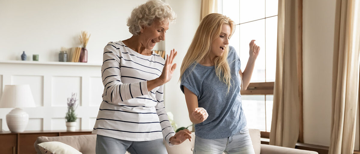 Zwei Frauen, eine in den 30ern und eine in den 50ern, tanzen ausgelassen in einem Wohnzimmer vor dem Sofa