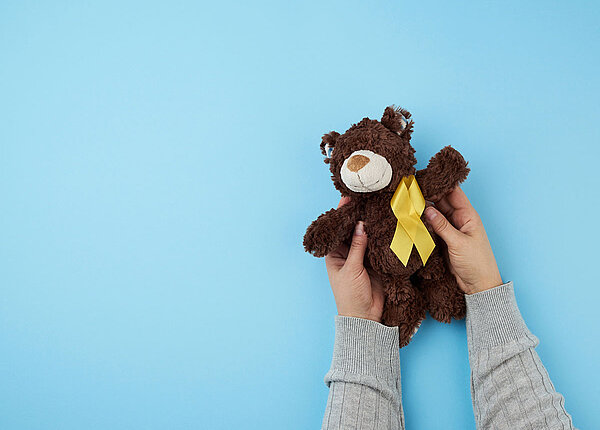 Kinderhände halten dunkelbraunen Teddybär vor blauem Hintergrund hoch. Auf der Brust des Bären ist eine gelbe Krebsschleife befestigt.