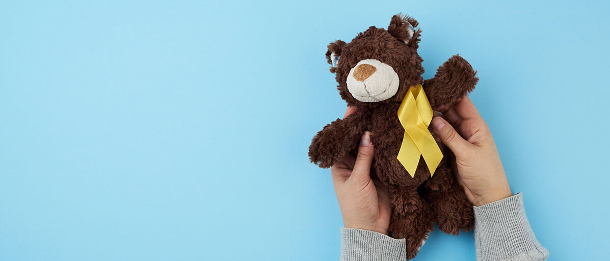 Kinderhände halten dunkelbraunen Teddybär vor blauem Hintergrund hoch. Auf der Brust des Bären ist eine gelbe Krebsschleife befestigt.