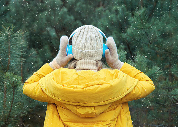 Hochsensibilität: Was sind Anzeichen bei hochsensiblen Menschen? Eine Frau in Winterkleidung wendet sich vom Betrachter ab und einem Nadelwald zu. Sie trägt Kopfhörer und hält diese zusätzlich fest.