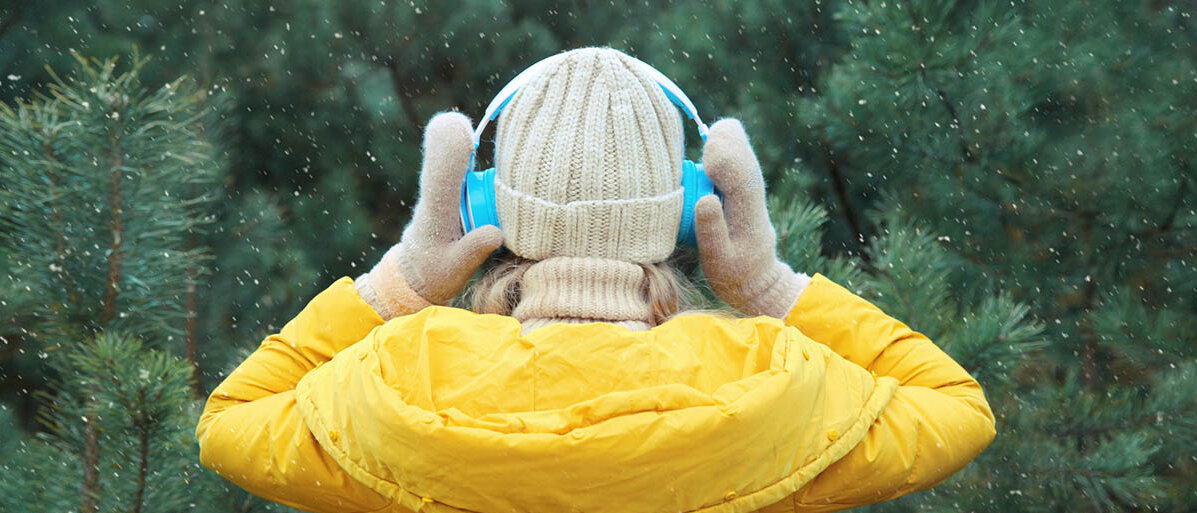 Hochsensibilität: Was sind Anzeichen bei hochsensiblen Menschen? Eine Frau in Winterkleidung wendet sich vom Betrachter ab und einem Nadelwald zu. Sie trägt Kopfhörer und hält diese zusätzlich fest.