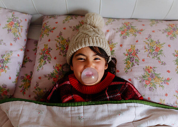 Ein Mädchen liegt mit Mütze im Bett und macht eine Kaugummi-Blase.