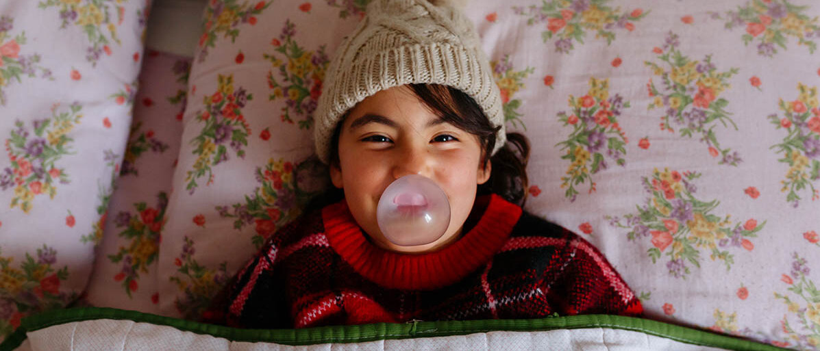 Kaugummi zur Grippe-Früherkennung: Selbsttest zur Influenza-Diagnose Ein Mädchen liegt mit Mütze im Bett und macht eine Kaugummi-Blase.