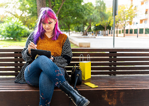 Hochsensibilität: Was sind Anzeichen bei hochsensiblen Menschen? Eine junge Frau mit pinken und violetten Haaren sitzt allein auf einer Parkbank und nutzt ein Tablet.