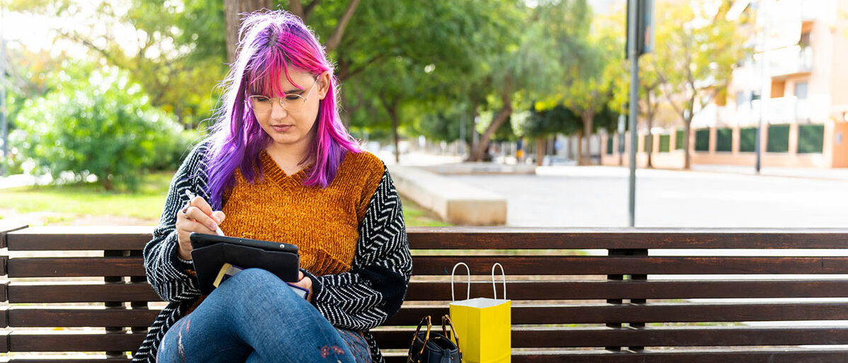 Hochsensibilität: Was sind Anzeichen bei hochsensiblen Menschen? Eine junge Frau mit pinken und violetten Haaren sitzt allein auf einer Parkbank und nutzt ein Tablet.