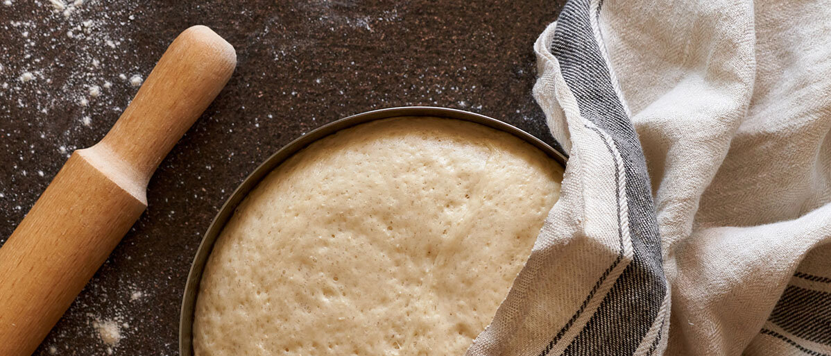 Roher Hefeteig in einer Schüssel leicht bedeckt von einem Küchenhandtuch. Nebendran liegt ein Nudelholz.