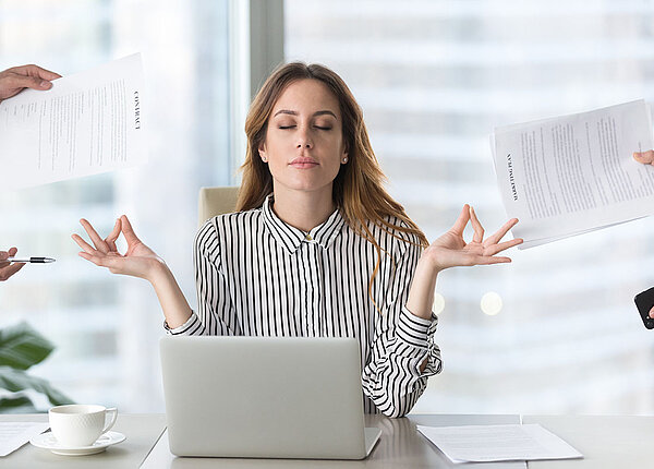 Frau sitzt vor ihrem Laptop und macht mit ihren beiden Händen eine meditative Bewegung. Von rechts und links werden Blätter von Kollegen hingehalten.