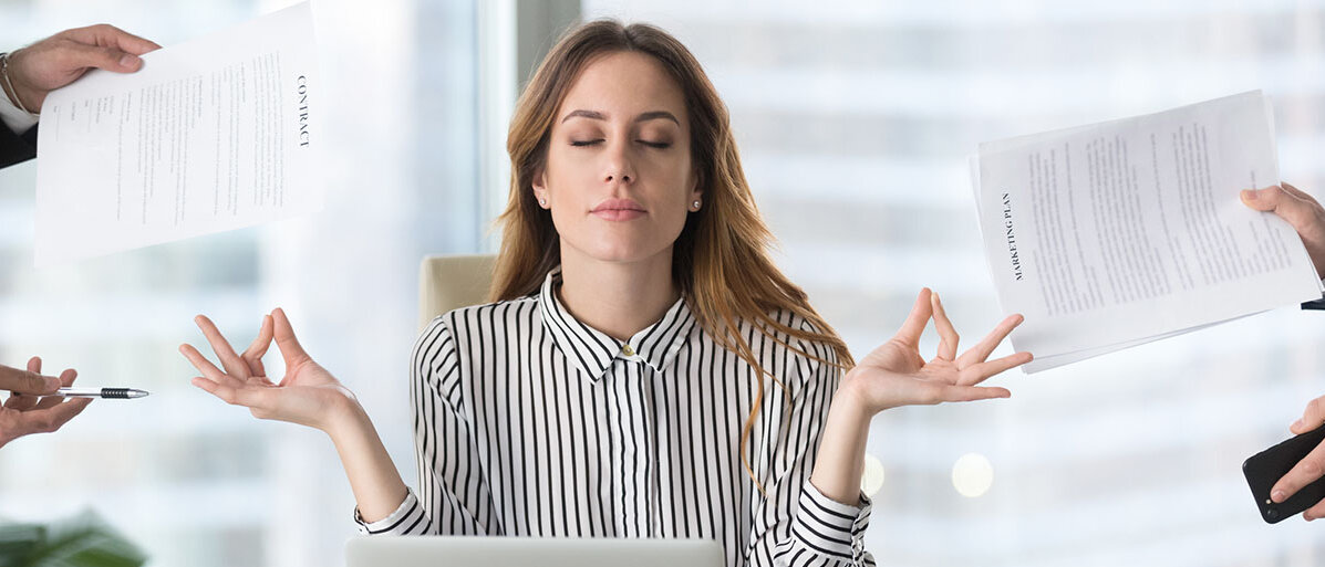 Multitasking im Arbeitsalltag vermeiden Frau sitzt vor ihrem Laptop und macht mit ihren beiden HÀnden eine meditative Bewegung. Von rechts und links werden BlÀtter von Kollegen hingehalten.