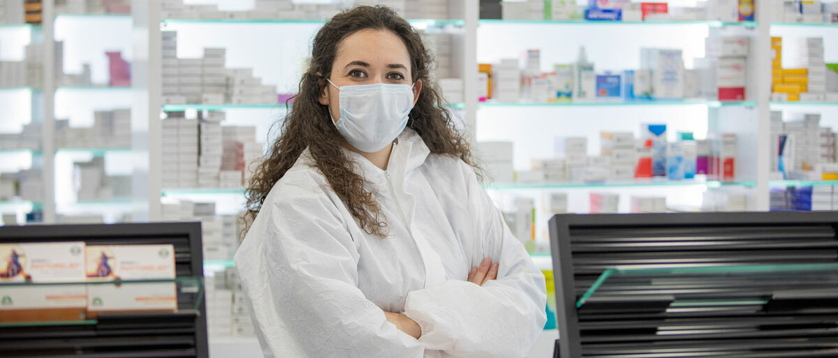 Eine Apothekenmitarbeiterin trägt Mundschutz und Kittel, sie steht mit verschränkten Armen hinter dem HV-Tisch.