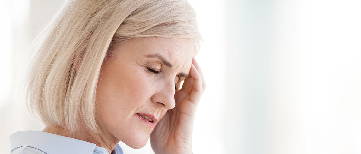 Frau mittleren Alters hält sich mit einer Hand die Schlfe und hat ein schmerzverzerrtes Gesicht.