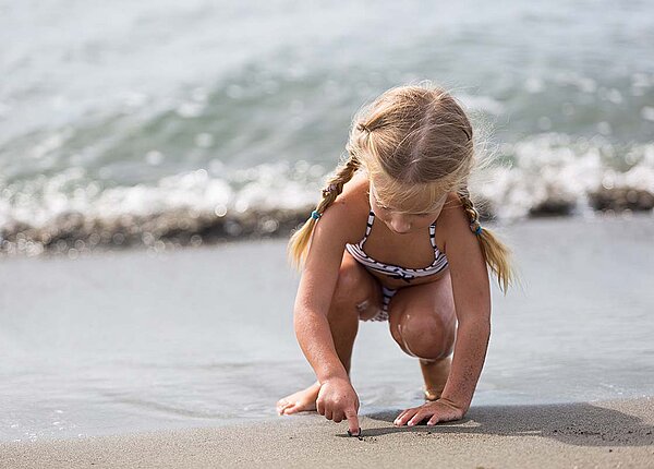 Portrait-Darstellung eines kleinen Mädchens, das einen schwarz-weiß gestreiften Bikini trägt, im Sand am Strand kniet und mit dem Finger etwas in den Sand malt.