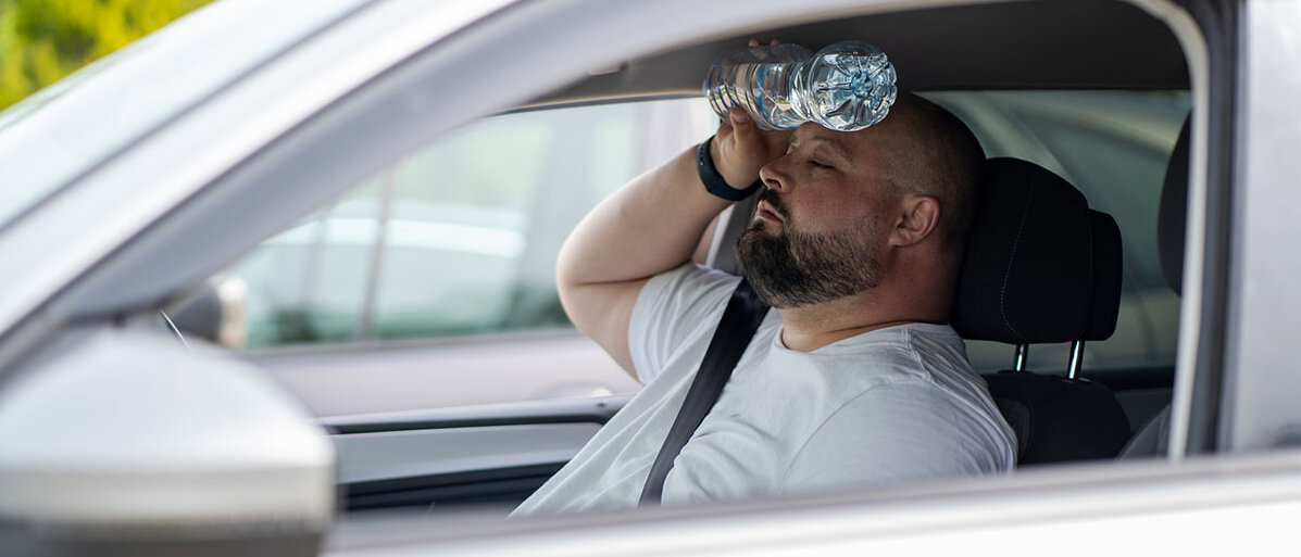 Ein Mann sitzt bei heruntergelassenen Fenstern im Auto und hält sich eine Wasserflasche an die Stirn.
