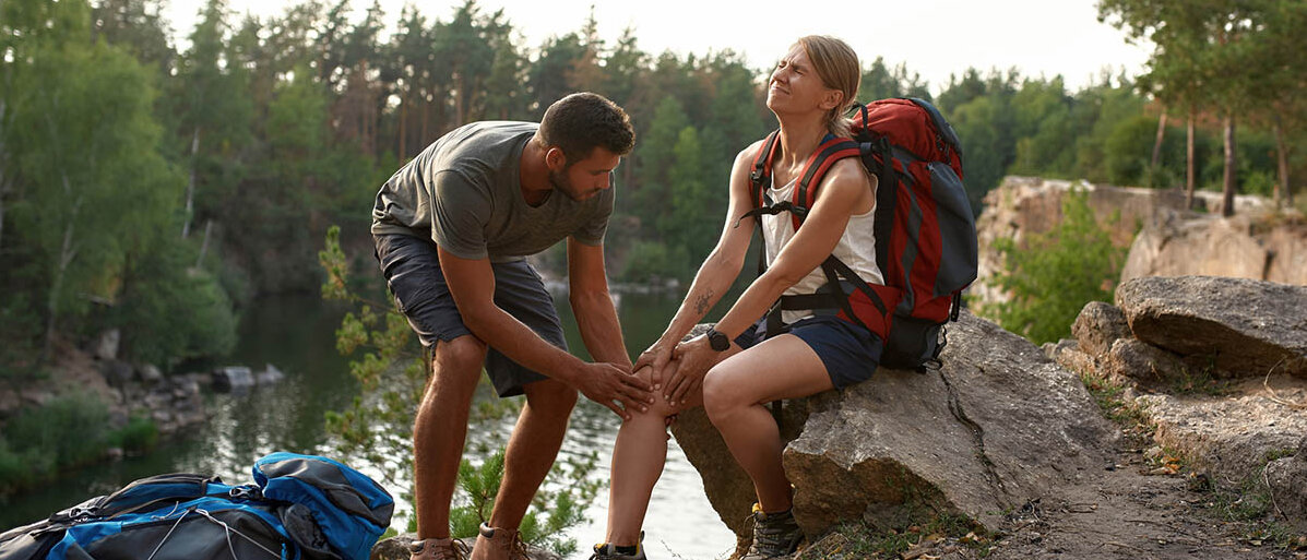 Ein Paar wandert. Sie sitzt auf einem Stein nahe einem Fluss, hält sich das Knie und verzieht das Gesicht vor Schmerz. Er hält ebenfalls ihr Knie und begutachtet es hilflos.