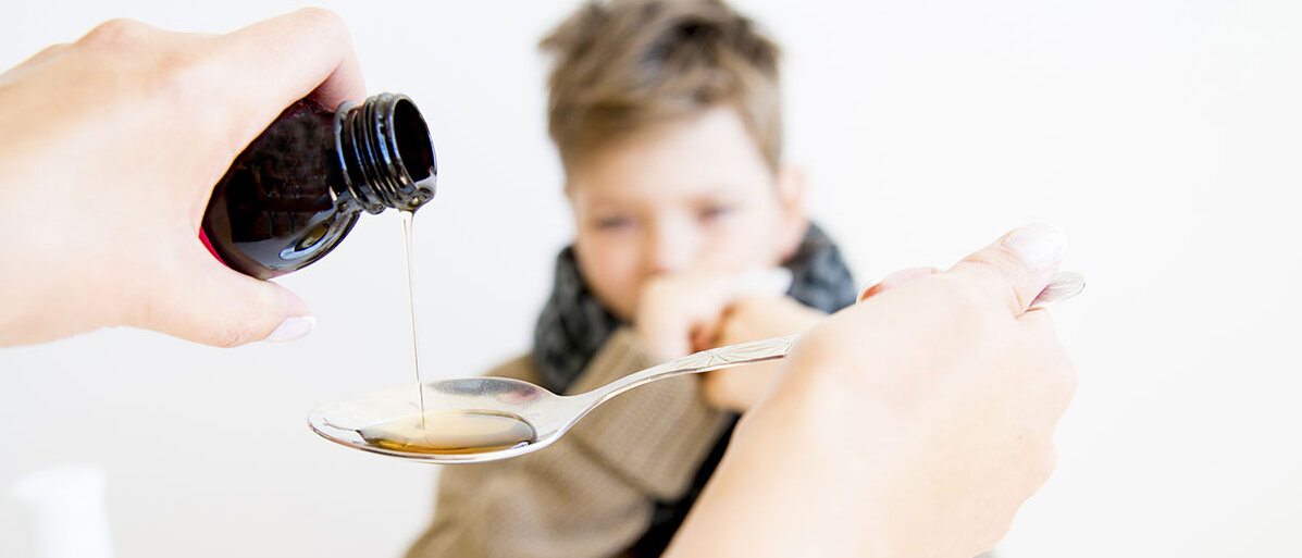 Ein kränkelnder Junge bekommt eine flüssige Arznei aus einem dunklen Fläschchen auf einem Esslöffel serviert. 