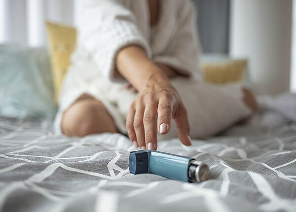 Eine Frau sitzt im Schneidersitz auf dem Bett und greift nach einem blauen Dosieraerosol.