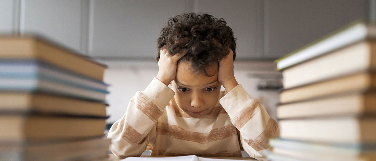 Schulstress bei Kindern Ăberforderter Junge schlĂ€ngt â umringt von BĂŒchern â HĂ€nde ĂŒber dem Kopf zusammen