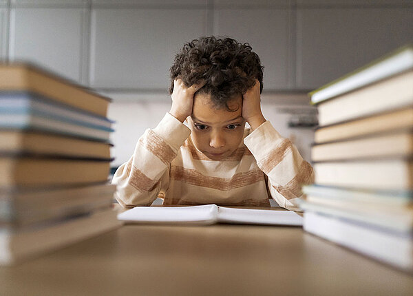 Schulstress bei Kindern Überforderter Junge schlängt – umringt von Büchern – Hände über dem Kopf zusammen