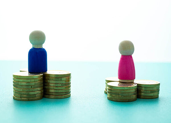 Gender Pay Gap bei den Gesundheitsberufen: Gehalt und Vedienst von Männern wesentlich höher als von Frauen Ein blaues Holzmännchen steht auf einem höheren Münzstapel als ein rosa Holzfigürchen.