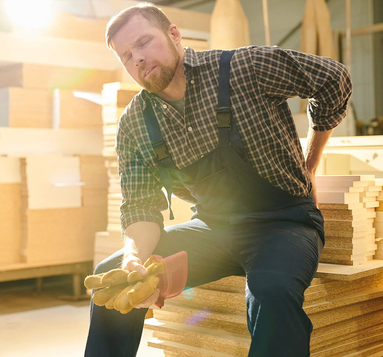 Ein Mann in Latzhose und Holzfällerhemd sitzt in einem Warenlager auf einem Stapel Kartons, beugt sich leicht nach vorne und hält sich den Lendenwirbelbereich. 