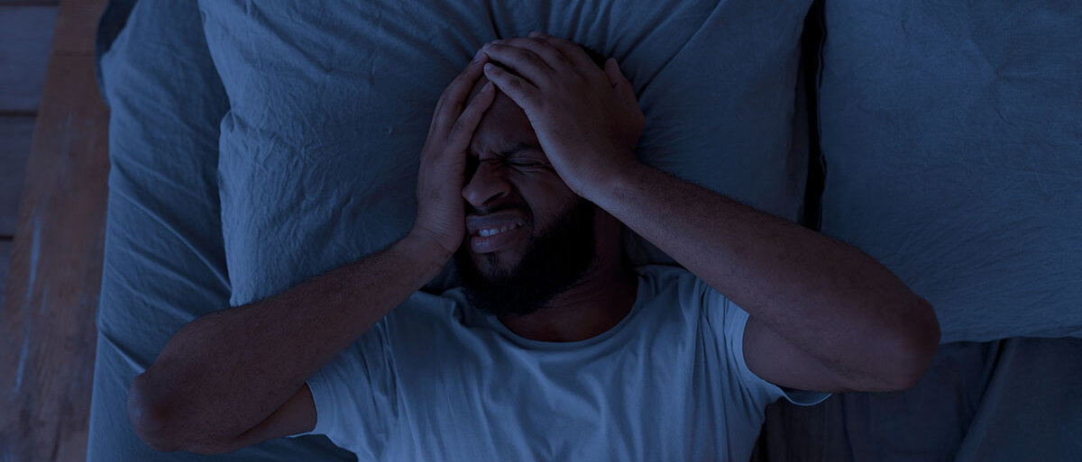 Ein Mann liegt im Dunkeln im Bett, fasst sich an den Kopf und verzieht das Gesicht.