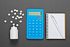 Auf einer grauen Unterlage liegt eine Dose, aus der Tabletten herausgerollt sind, ein blauer Taschenrechner sowie ein Karoblock mit Stift.