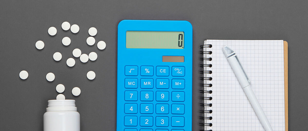 Auf einer grauen Unterlage liegt eine Dose, aus der Tabletten herausgerollt sind, ein blauer Taschenrechner sowie ein Karoblock mit Stift.