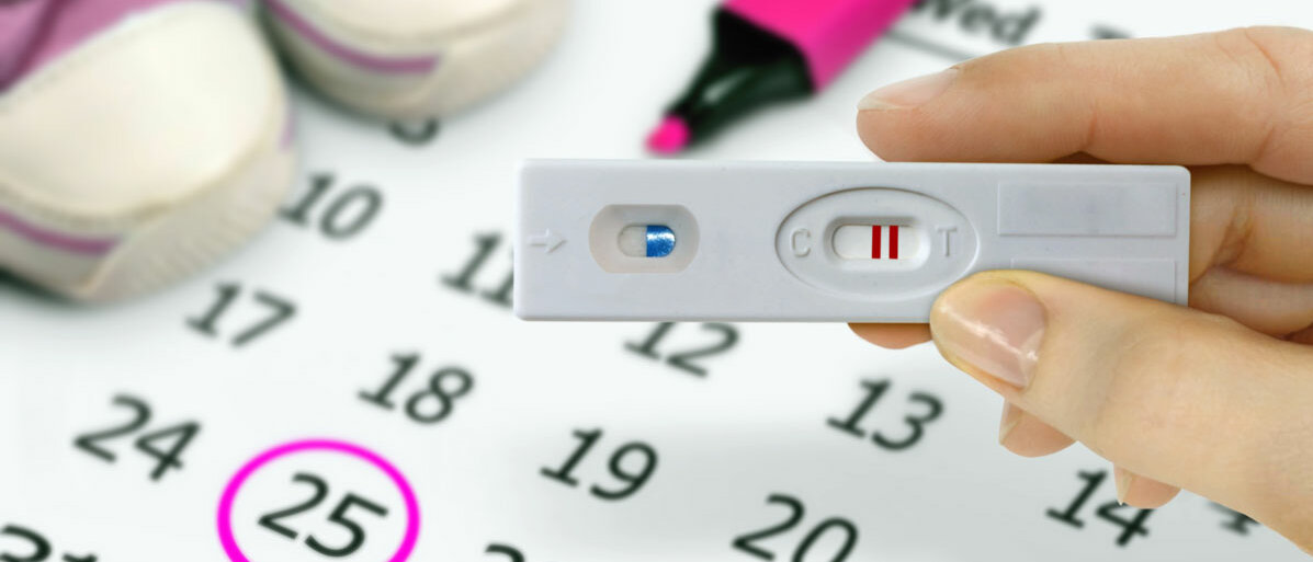 Auf einem Kalender ist die Zahl 25 eingekreist. Darauf stehen ein Paar Babyschuhe. Eine Hand hält einen positiven Schwangerschaftstest.